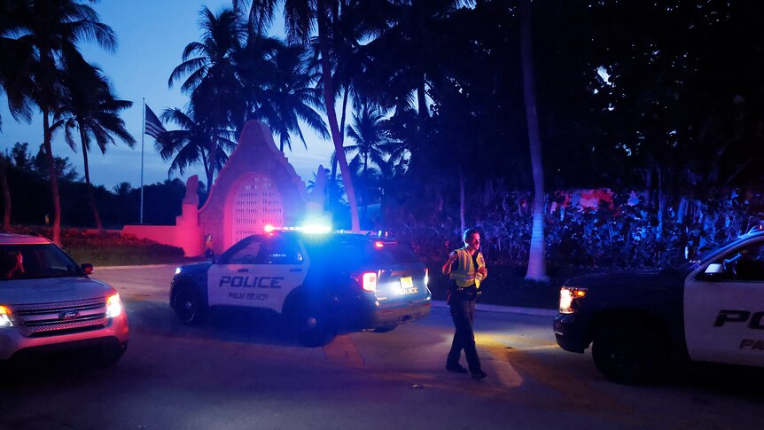 A police officer stands next to a lit-up squad car outside a mansion at dusk, surrounded by palm trees 