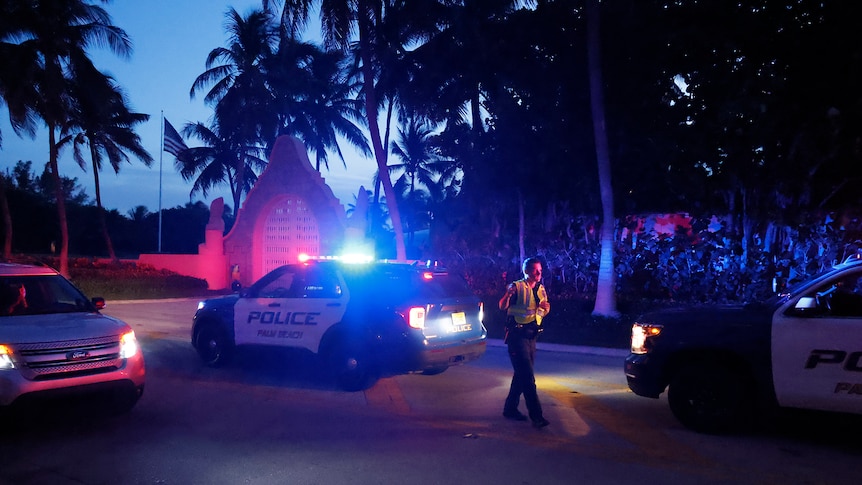 A police officer stands next to a lit-up squad car outside a mansion at dusk, surrounded by palm trees 