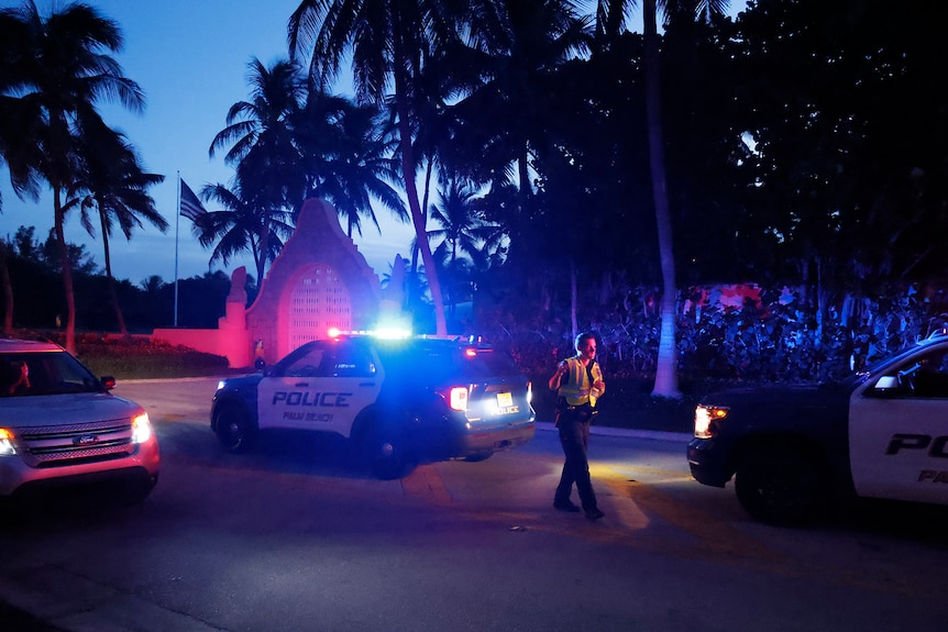 A police officer stands next to a lit-up squad car outside a mansion at dusk, surrounded by palm trees 