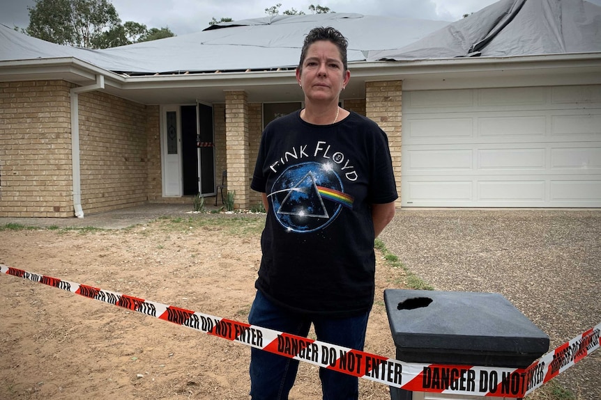 A woman wearing a pink floyd shirt standing outside a single storey house with a tarp covering the roof