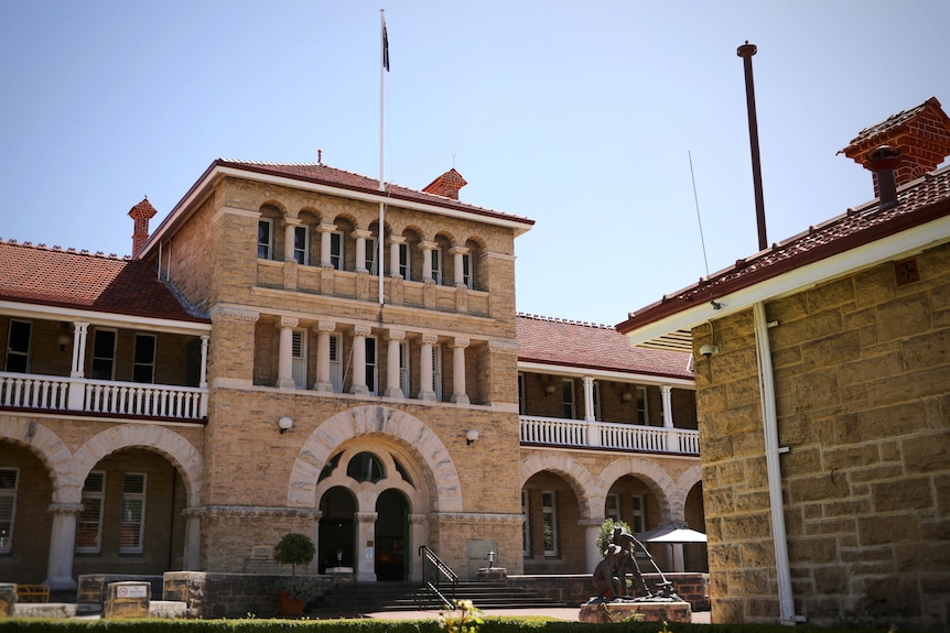 A historic three-storey building with arches and columns and a flag hanging limply from a pole. It has a statue in front of it