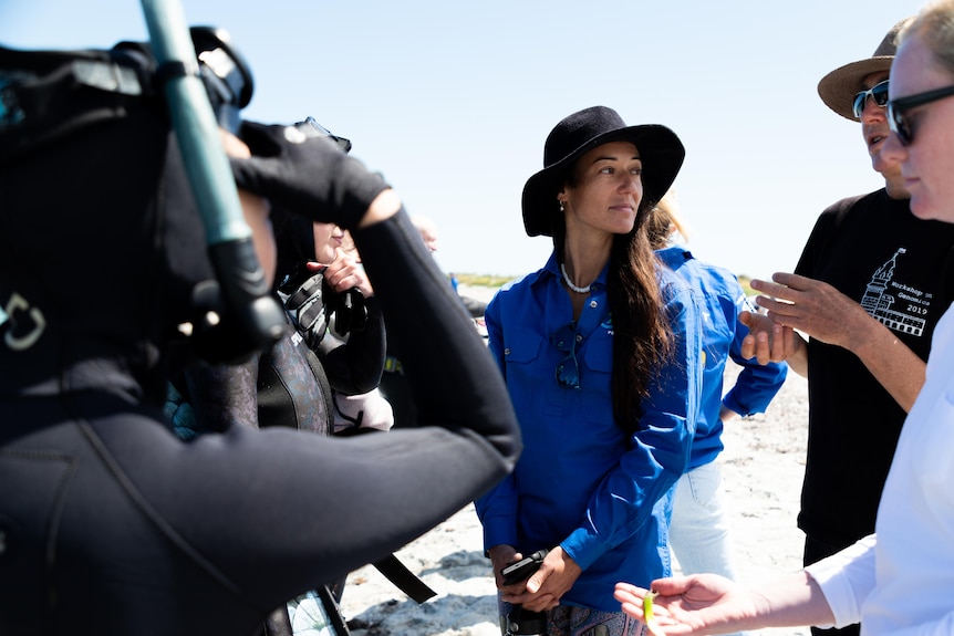 A woman in a wide-brim hat and blue shirt stands in a crowd of people wear wet suits on the beach.