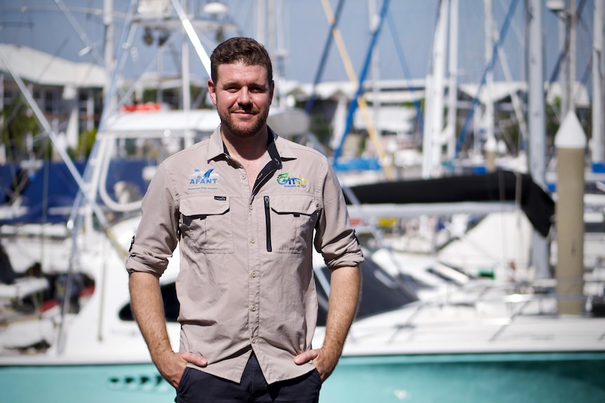 A man in a khaki collared shirt standing in front of boats moored at a marina, on a sunny day.
