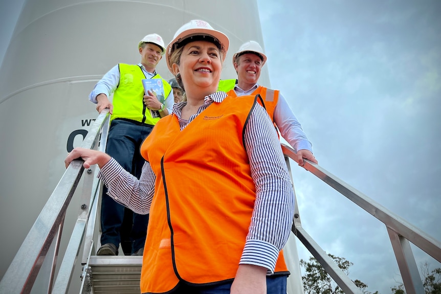 The Premier wearing an orange hi-vis vest stands on a steel ladder in front of two men also wearing hi-vis and hard hats.