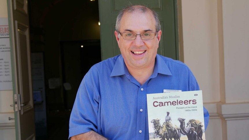 A man with glasses standing in front of a door holding a book called Australia's Muslim cameleers