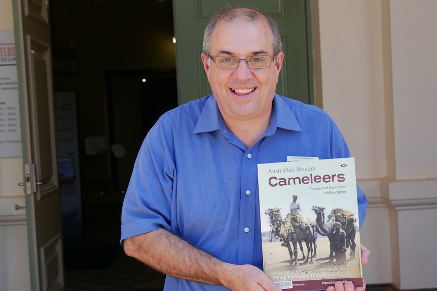 A man with glasses standing in front of a door holding a book called Australia's Muslim cameleers