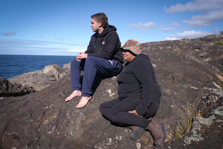 A serious young man and older woman sitting on large rock by the ocean.