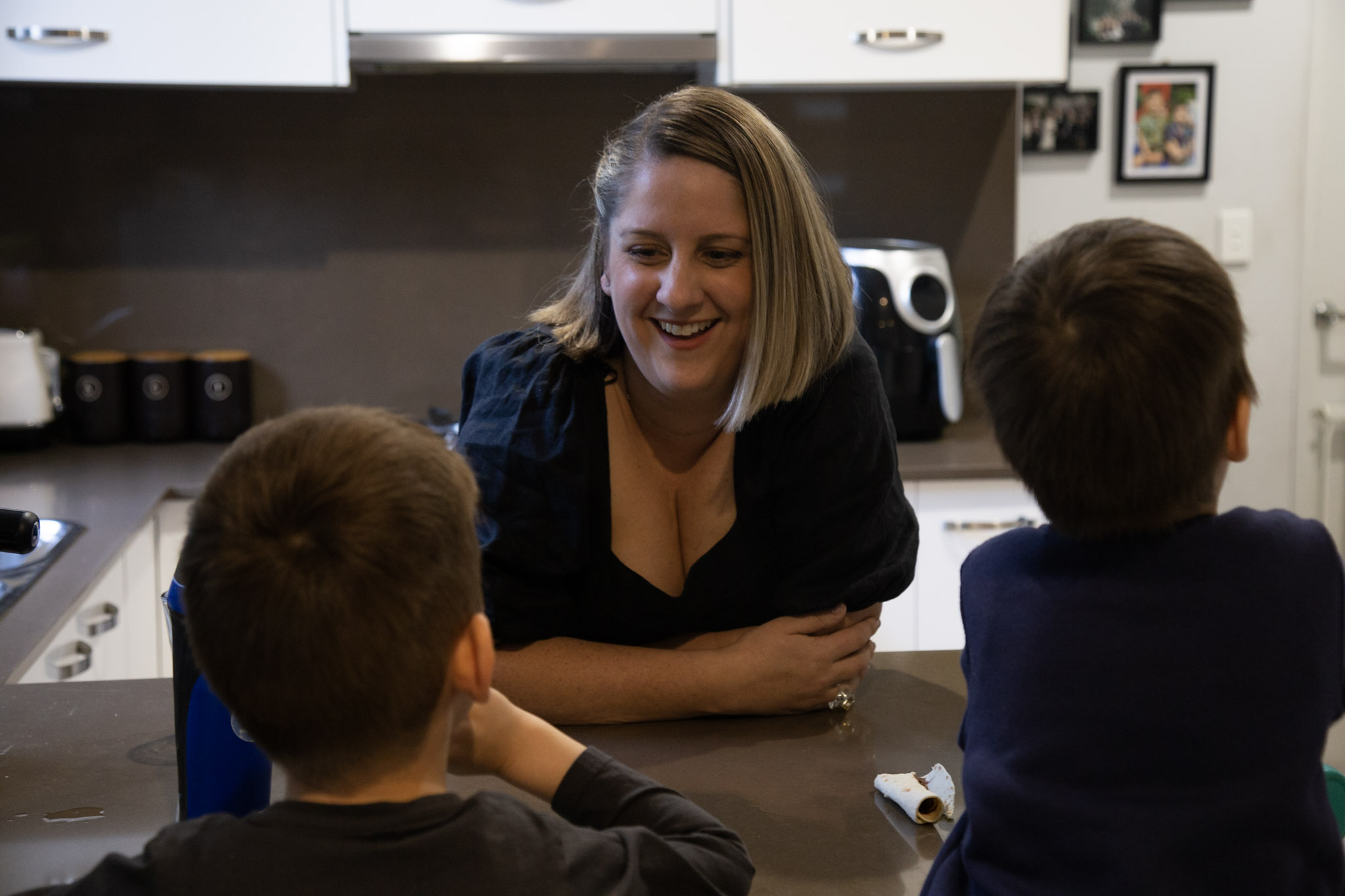 Woman stands leaning over kitchen bench laughing with two young boys.