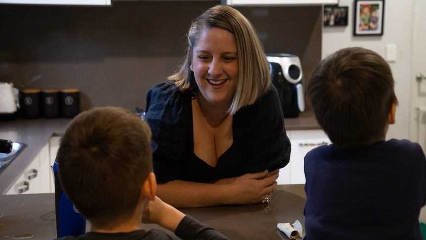 Woman stands leaning over kitchen bench laughing with two young boys.