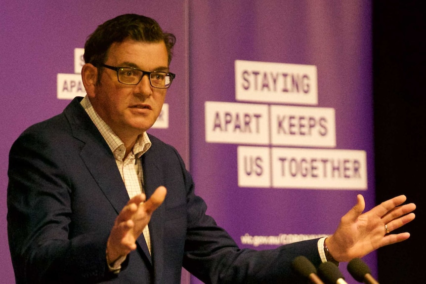 Daniel Andrews wearing a jacket, shirt but no tie stands at a podium with his arms out in front of him.