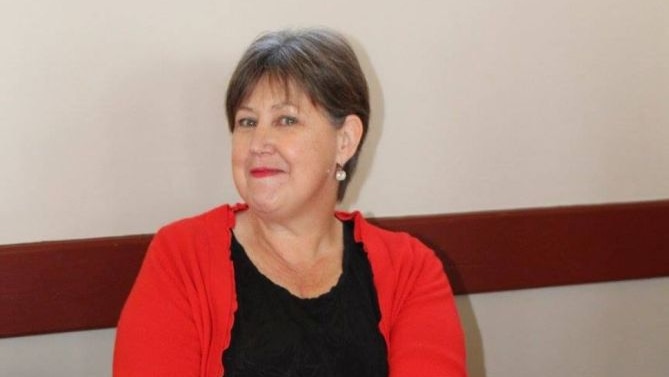 Lady in a red cardigan, seated against a wall.