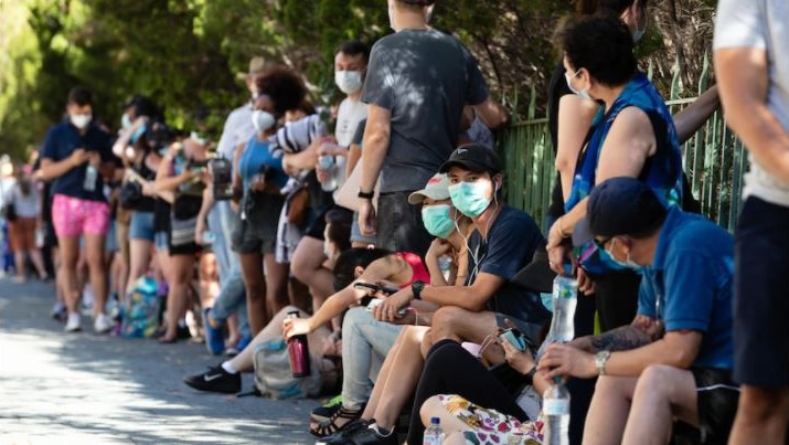 Lines of people waiting along the side of the street for a covid test, many sitting and standing wearing masks.