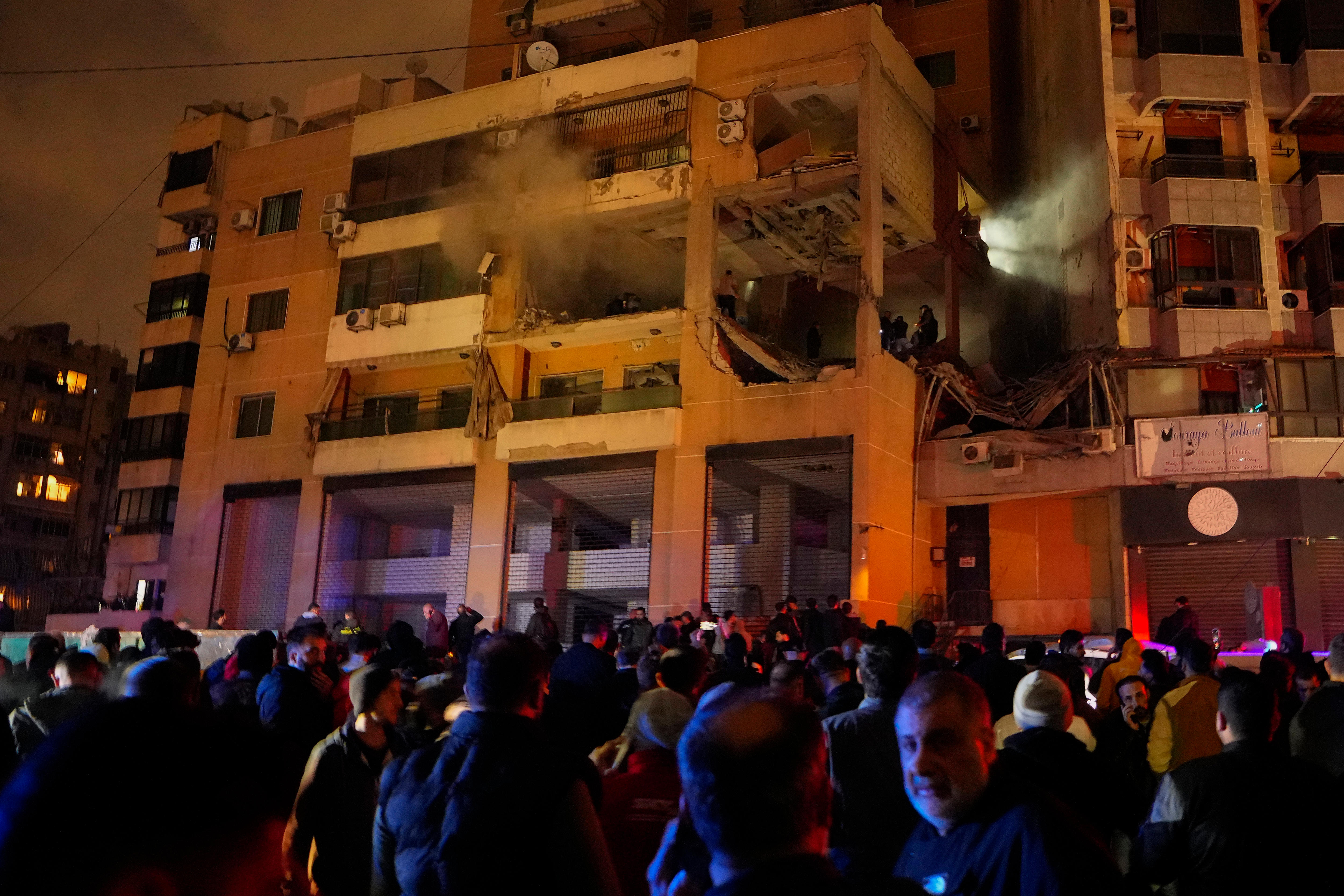 A crowd around an apartment building that is damaged with smoke coming out. 