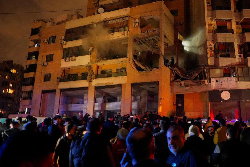 A crowd around an apartment builidng that is damaged with smoke coming out. 