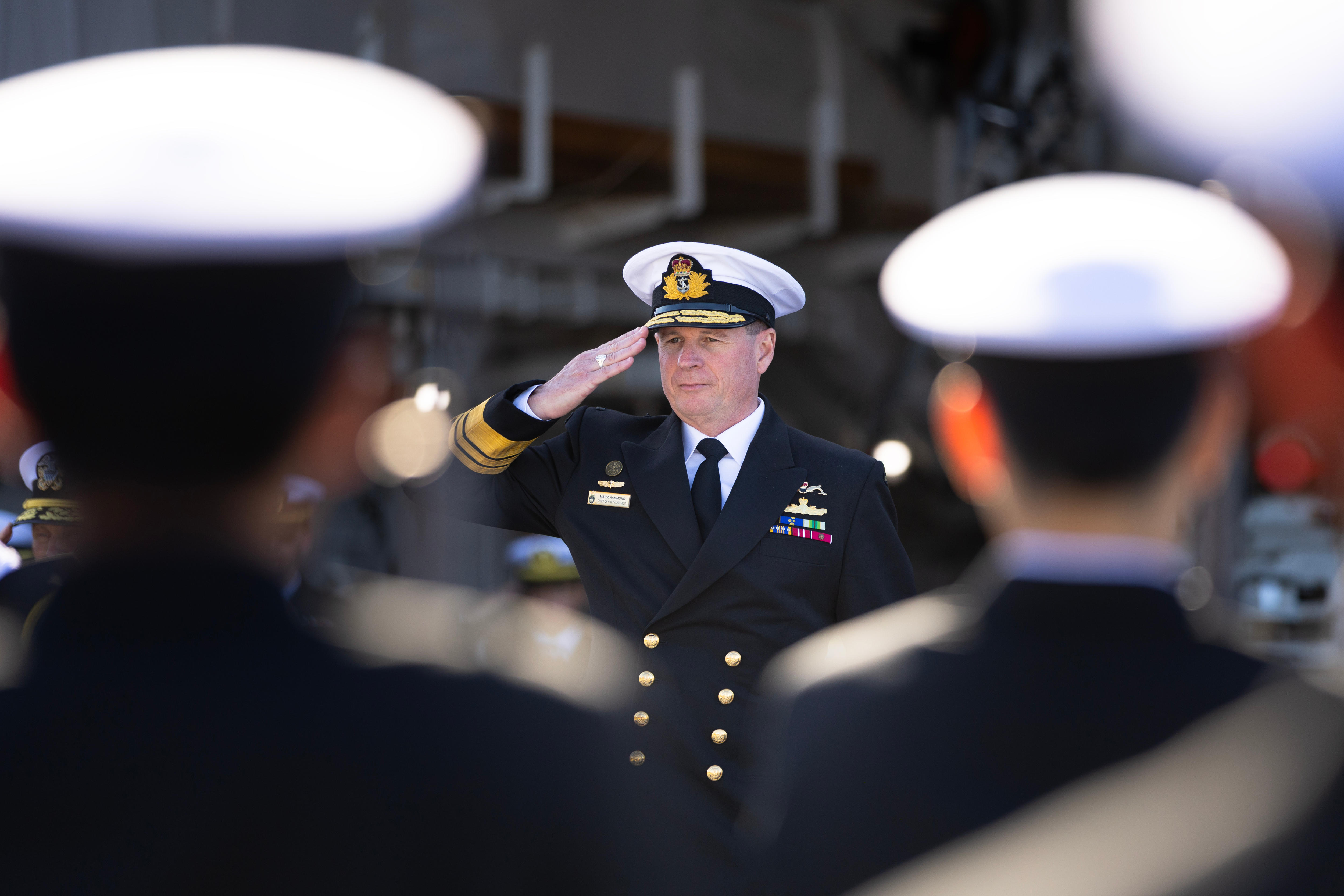 A middle-aged man in an Australian navy uniform salutes a group of sailors.