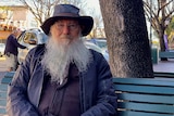 A man with a wide brim hat and a large beard sits on a public bench.