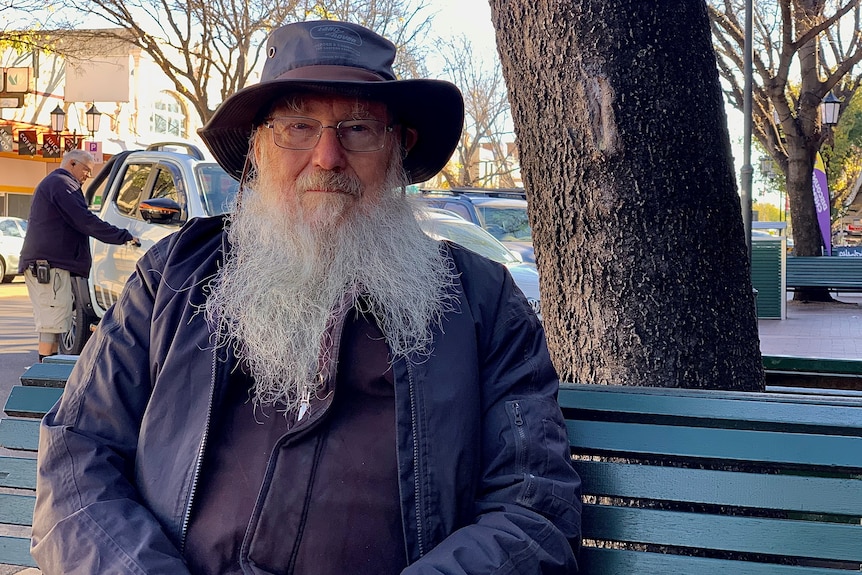 A man with a wide brim hat and a large beard sits on a public bench.