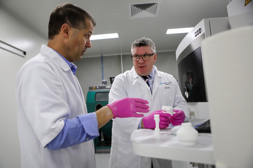 Man in white coat and blue mask holds up  small set of white tubes wearing thick white gloves in a lab type setting