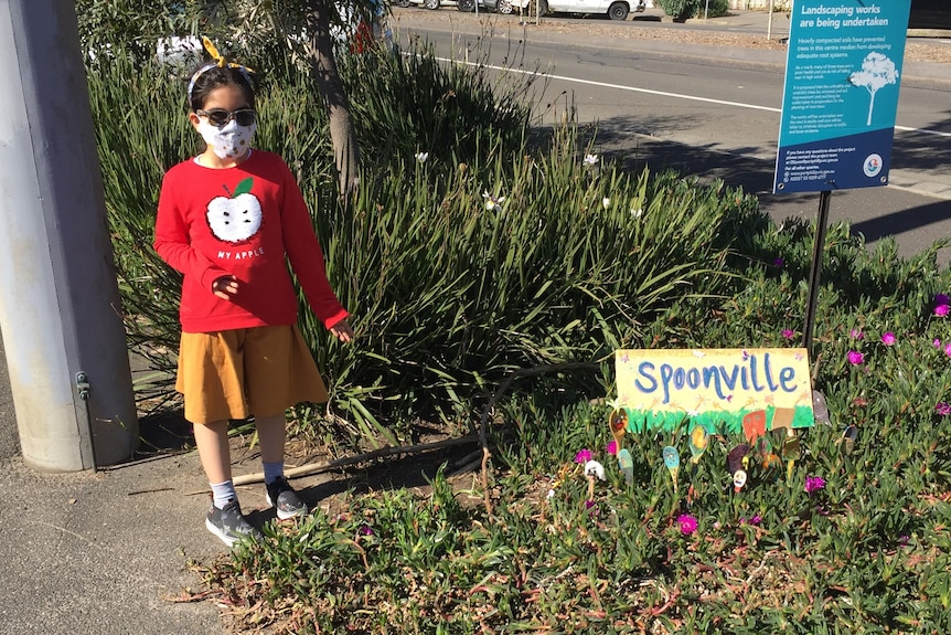 A girl in sunglasses and a mask stands next to an artistic village of spoons dubbed 'Spoonville'.