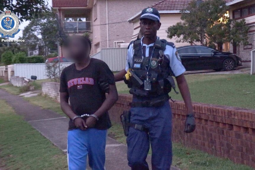 A man, with his face blurred, is in handcuffs as a police man takes him towards a car