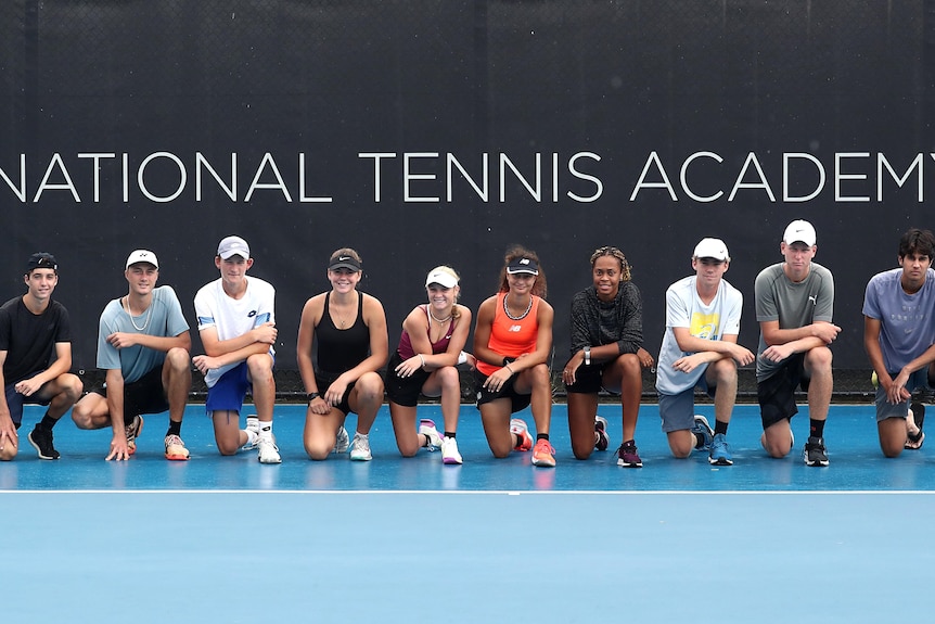 A group of tennis players kneelfor a photo in front of a sign that says NATIONAL TENNIS ACADEMY