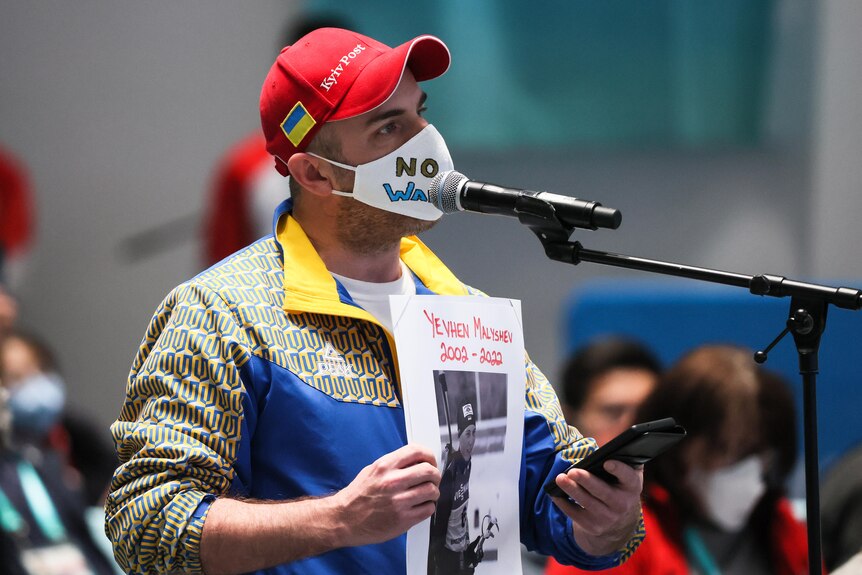 A man wearing a mask saying "no war in UKR" stands at a microphone holding a picture saying "Yevhen Malshev 2002-2022".