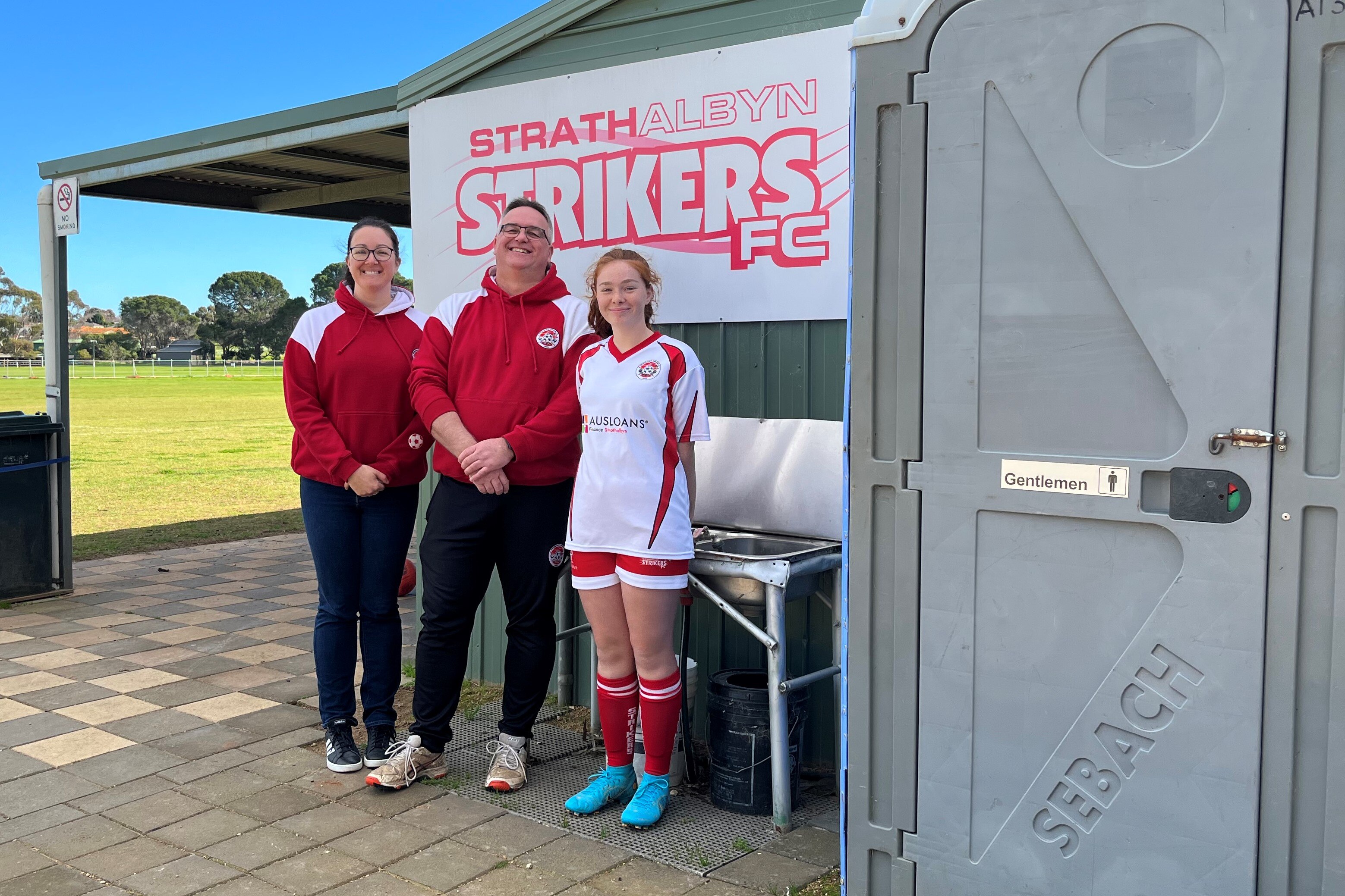 Two adults and a young player stand next to a shed and a gate