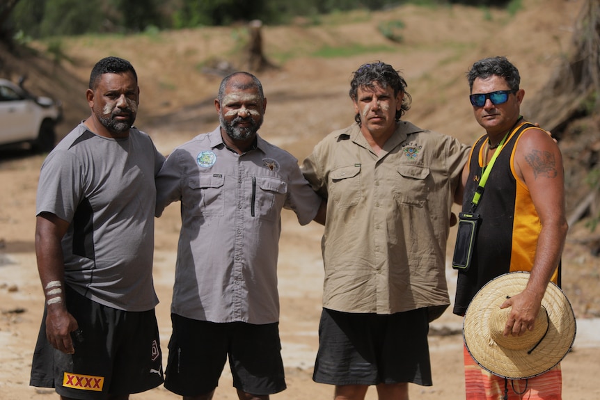 Four Indigenous men stand with their arms around each other - three of them have face paint.
