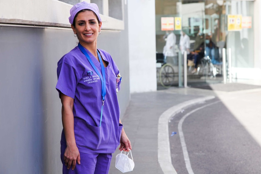 A woman in purple scrubs stands outside a hospital entrance with a face mask in her hand