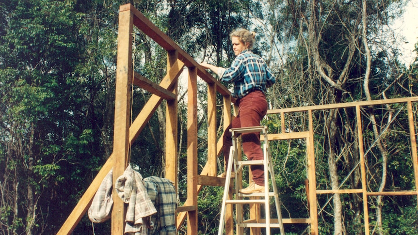 Medium colour archive shot of a woman standing on a ladder while building a timber hut.