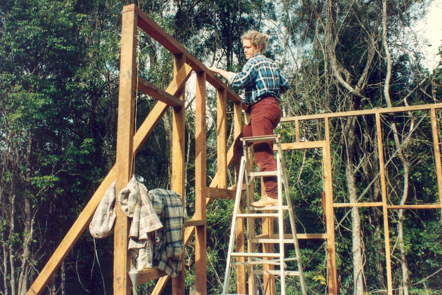 Medium colour archive shot of a woman standing on a ladder while building a timber hut.