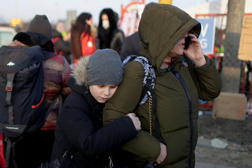 A person wearing a hooded green jacket talks on the phone while a young child wearing a grey beanie holds onto their arm.