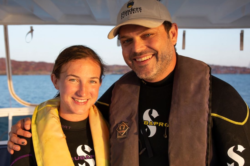 A woman in a diving suit with a floatation device around her neck is embraced by a man in similar suit wearing a university cap