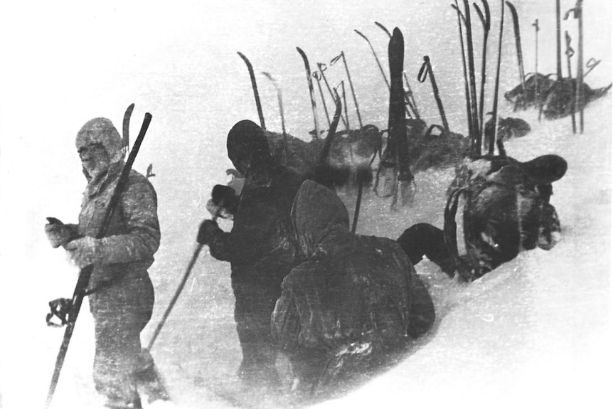 A black and white photo of several skiers in deep snow with polls and skis sticking into the air 
