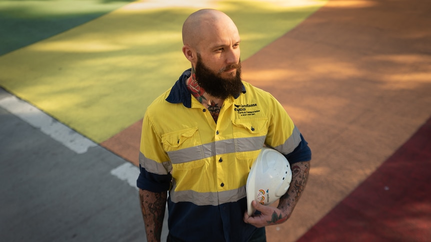 A bald man wearing hi-vis and carrying a white hard hat looks away to his left