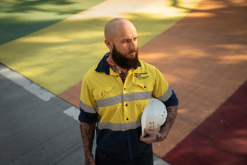A bald man wearing hi-vis and carrying a white hard hat looks away to his left