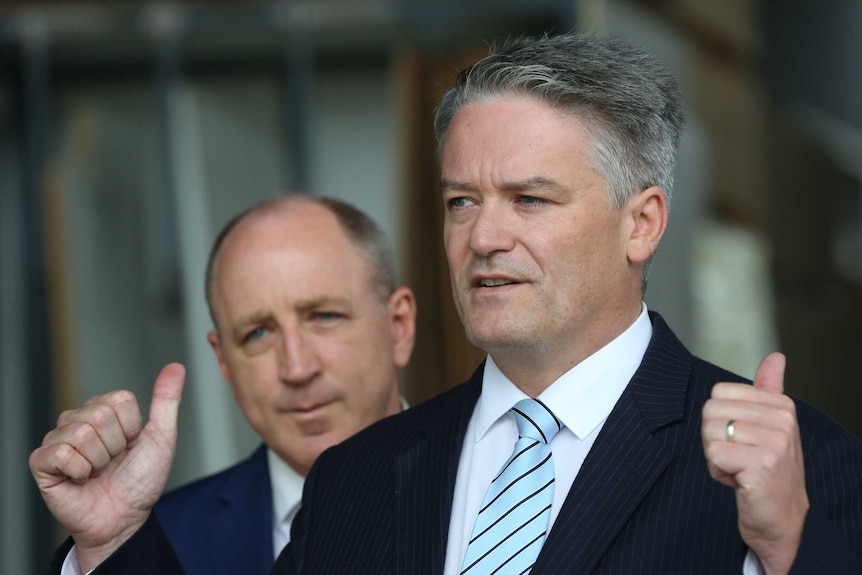 Mathias Cormann holds up two thumbs in a press conference. He's wearing a light blue striped tie. Luke Howarth is behind him.