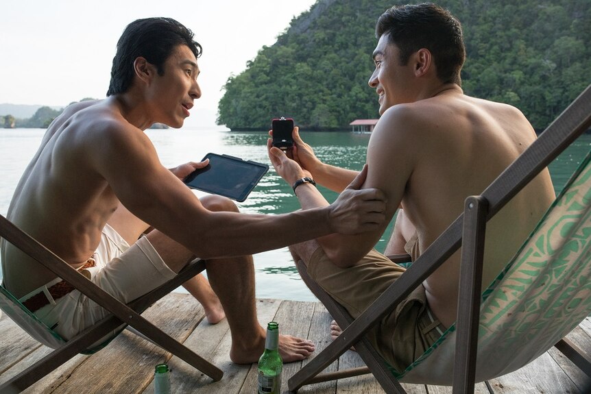 Stars Chris Pang and Henry Golding sitting by a river with Golding revealing a wedding ring, a still from Crazy Rich Asians.