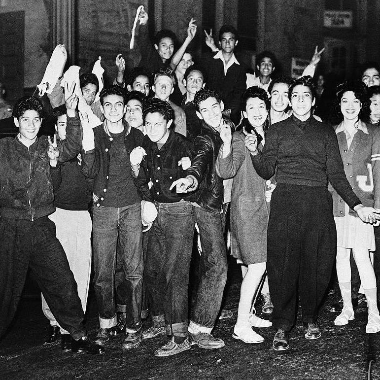 A group of youths gather for a group photo, some of them are wearing leather jackets and zoot suits. They are smiling and posing