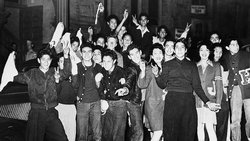 A group of youths gather for a group photo, some of them are wearing leather jackets and zoot suits. They are smiling and posing