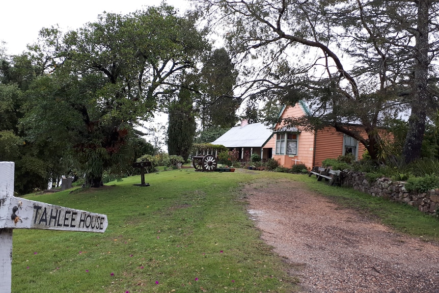 An old homestead, at the end of a driveway, surrounded by green lawns with a sign saying "Tahlee House".