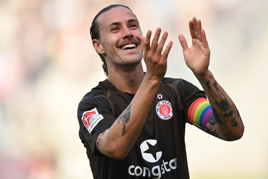 A male soccer player wearing a brown shirt and rainbow armband applauds