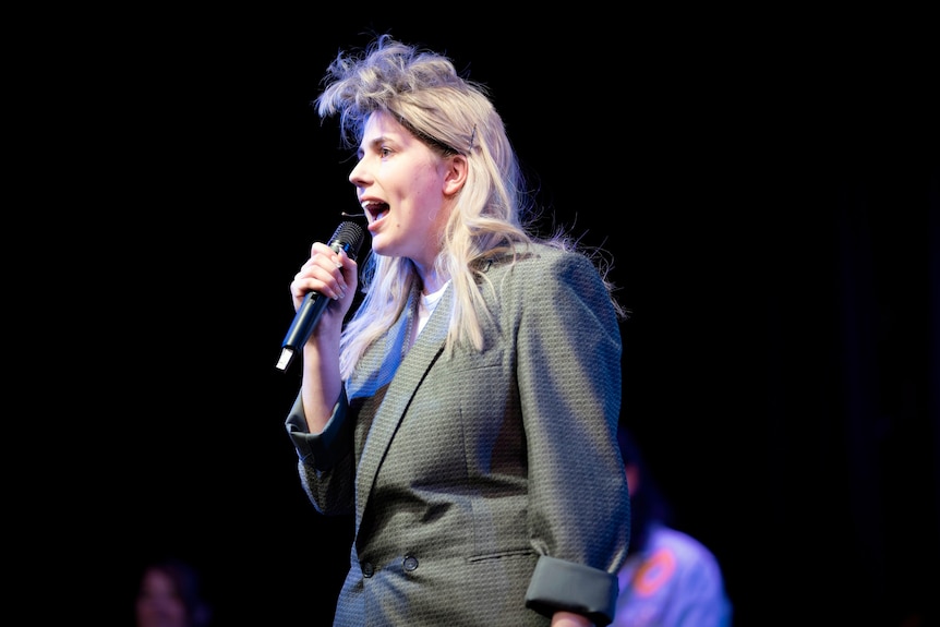 A teenage girl wearing a blonde mullet wig sings into a microphone.