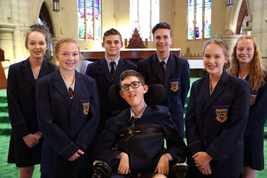 Seven students in their school uniform pose for a photo. Thomas Byrne centre in wheelchair. Chapel stainglass windows in back