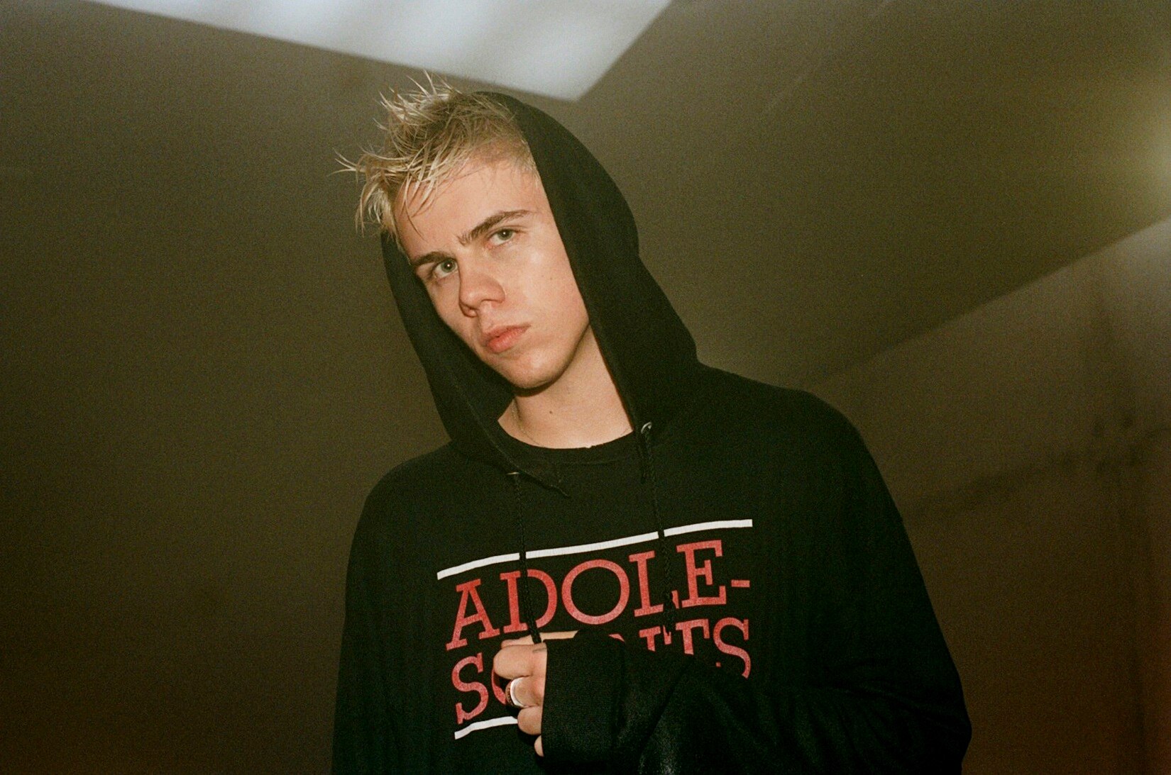 The Kid LAROI stands looking down at the camera in a black Adolescents hoodie with the hood up over his spiked blonde hair