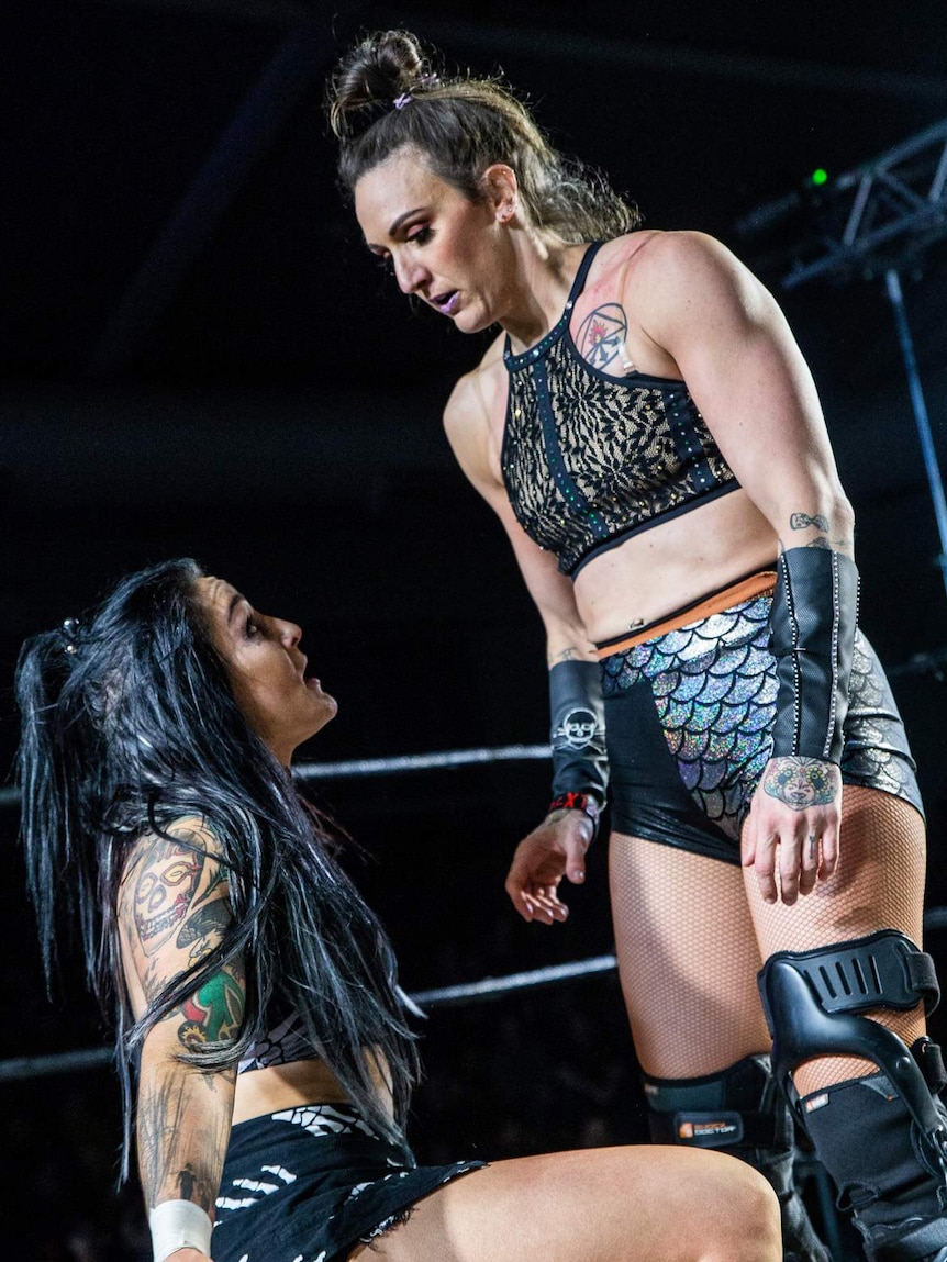 Female wrestler in silver shorts leaning over another female wrestler with long black hair in the ring.