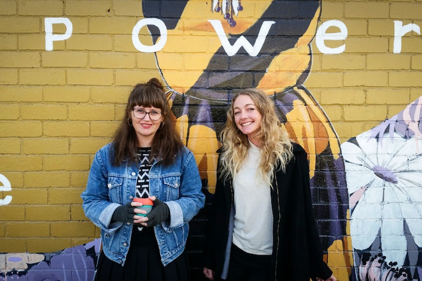 Two women stand smiling in front of a wall painted with a colourful mural.