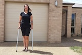 Woman standing with crutches on driveway in front of home looks at camera.