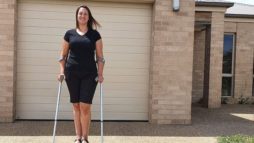 Woman standing with crutches on driveway in front of home looks at camera.