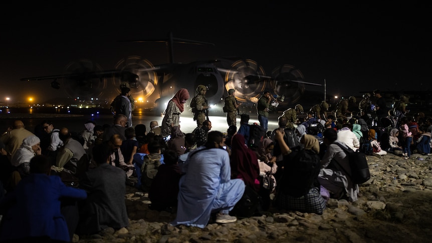 A group of Afghan evacuees sit on the ground with a large plane in the background. 